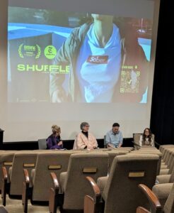 A distant picture of the 4 person discussion panel at the viewing of the documentary "Shuffle" with a frame of the movie behind them.