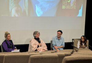 A picture of the 4 person discussion panel at the viewing of the documentary "Shuffle" with a frame of the movie behind them.